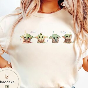 May include: Cream-colored sweatshirt featuring four cartoon Grogu characters in various poses. Each Grogu is green with large eyes and is surrounded by small stars and a ring. The baocake logo is visible in the lower left corner.