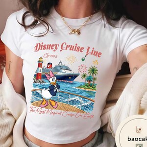 K&ouml;nnte beinhalten: Wei&szlig;es T-Shirt mit einem farbenfrohen Disney Cruise Line-Motiv. Das Design zeigt ein Kreuzfahrtschiff, einen Leuchtturm und Daisy Duck. Der Text lautet "Disney Cruise Line Est. 1998" und "The Most Magical Cruise On Earth".