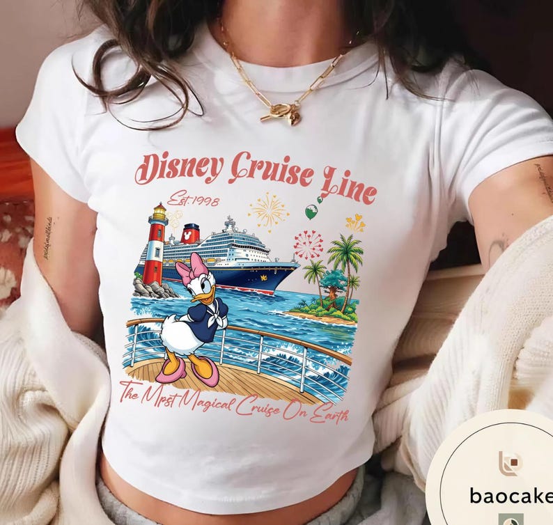 K&ouml;nnte beinhalten: Wei&szlig;es T-Shirt mit einem farbenfrohen Disney Cruise Line-Motiv. Das Design zeigt ein Kreuzfahrtschiff, einen Leuchtturm und Daisy Duck. Der Text lautet "Disney Cruise Line Est. 1998" und "The Most Magical Cruise On Earth".