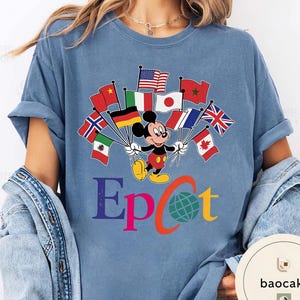 May include: A light blue t-shirt with a graphic of Mickey Mouse holding flags from different nations. The word "Epcot" is printed below in various colours, with a globe graphic. The shirt is made of a soft material.