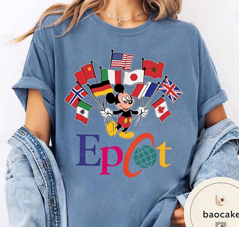 May include: A light blue t-shirt with a graphic of Mickey Mouse holding flags from different nations. The word "Epcot" is printed below in various colours, with a globe graphic. The shirt is made of a soft material.