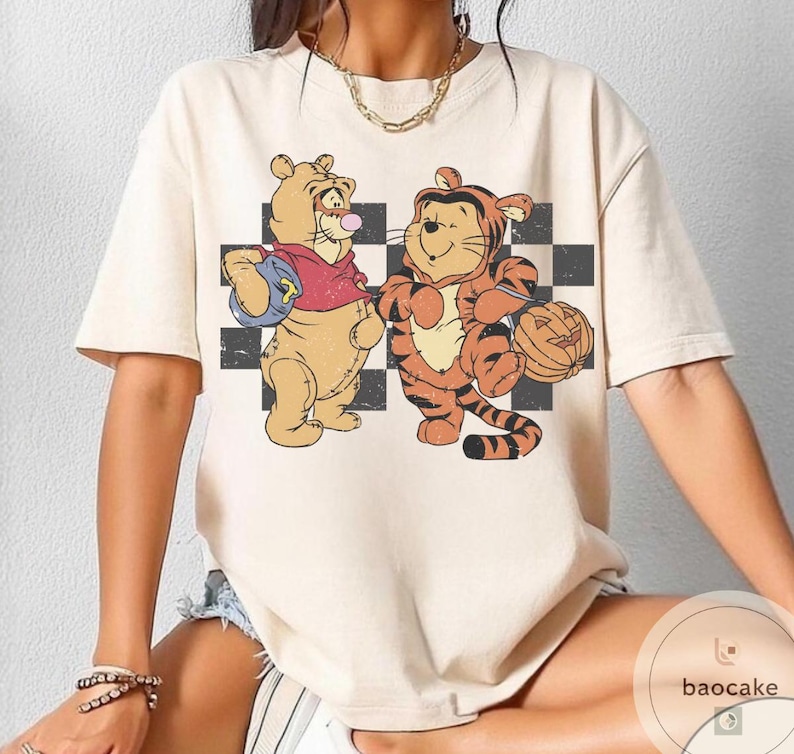 May include: Cream-colored t-shirt featuring a vintage-style illustration of Winnie the Pooh and Tigger dressed in Halloween costumes. Pooh wears a red shirt and carries a blue bag, while Tigger holds a carved pumpkin. The background has a black and white checkered pattern.