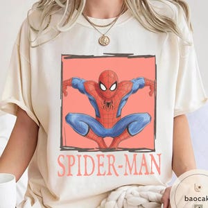 May include: Cream-colored t-shirt featuring a graphic of Spider-Man in a crouching pose, set against a coral background within a black outlined rectangle. The words "SPIDER-MAN" are printed in coral below the image. The superhero is depicted in his classic red and blue suit.