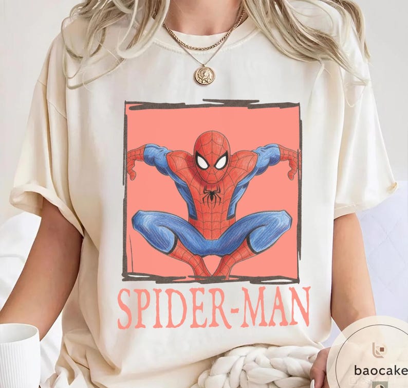 May include: Cream-colored t-shirt featuring a graphic of Spider-Man in a crouching pose, set against a coral background within a black outlined rectangle. The words "SPIDER-MAN" are printed in coral below the image. The superhero is depicted in his classic red and blue suit.