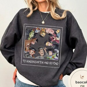 May include: Dark grey sweatshirt with a graphic of animated characters wearing graduation caps. The image includes characters from a popular animated movie. The text "TO KINDERGARTEN AND BEYOND" is printed in white.