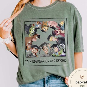 May include: Sage green t-shirt with a framed graphic of animated characters wearing graduation caps. The graphic includes the text "TO KINDERGARTEN AND BEYOND" in a black font. The shirt is made of a soft material.