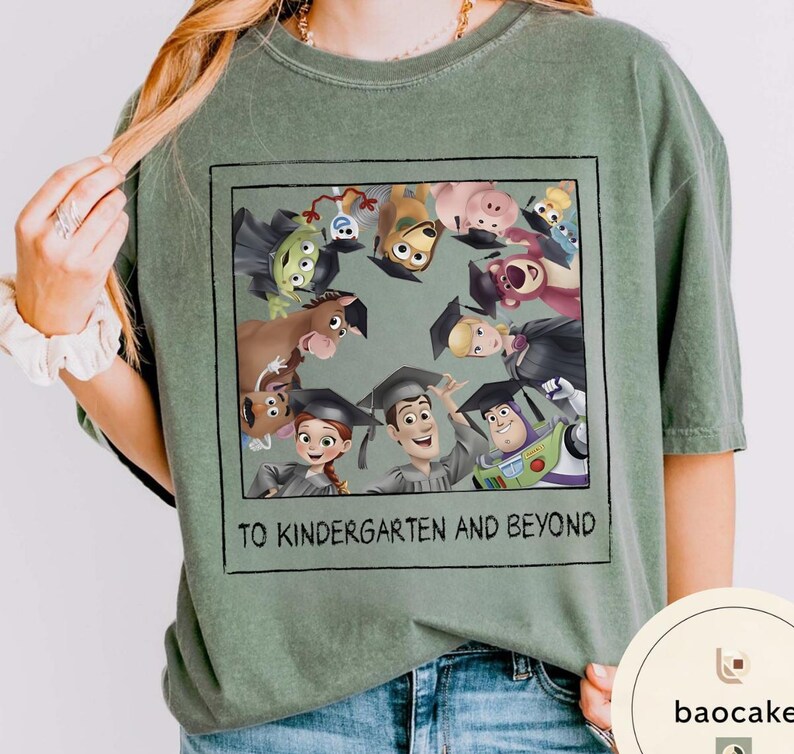 May include: Sage green t-shirt with a framed graphic of animated characters wearing graduation caps. The graphic includes the text "TO KINDERGARTEN AND BEYOND" in a black font. The shirt is made of a soft material.