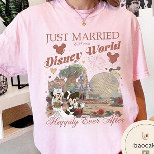 May include: A light pink t-shirt with a graphic featuring Mickey and Minnie Mouse in wedding attire, the words "Just Married Disney World" and "Happily Ever After". The design includes iconic Disney World landmarks and Mickey Mouse balloons.