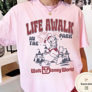May include: A light pink t-shirt featuring a graphic of Donald Duck running in front of a castle with fireworks. The text "LIFE AWALK IN THE PARK" and "Walt Disney World" are printed in burgundy.