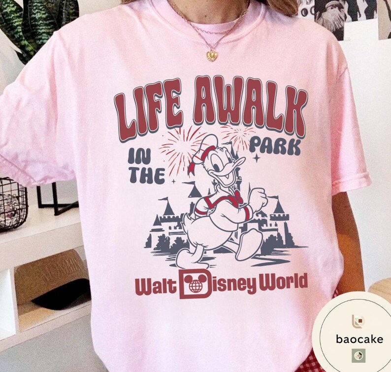 May include: A light pink t-shirt featuring a graphic of Donald Duck running in front of a castle with fireworks. The text "LIFE AWALK IN THE PARK" and "Walt Disney World" are printed in burgundy.