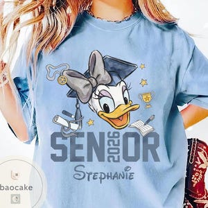 May include: Light blue t-shirt featuring a cartoon Daisy Duck wearing a graduation cap and bow. The shirt has the text "SENIOR 2026" and the name "Stephanie" in a script font. Includes stars, a trophy, and a book.