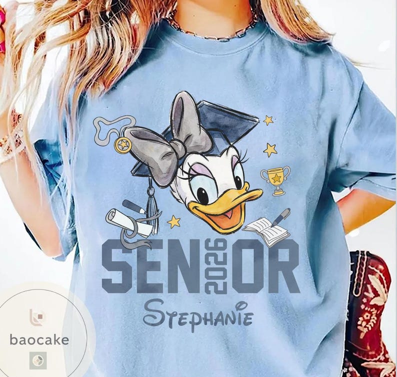 May include: Light blue t-shirt featuring a cartoon Daisy Duck wearing a graduation cap and bow. The shirt has the text "SENIOR 2026" and the name "Stephanie" in a script font. Includes stars, a trophy, and a book.