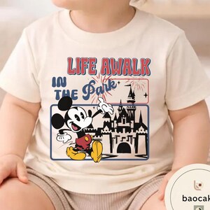 May include: Cream-coloured t-shirt featuring a graphic of Mickey Mouse with the text "LIFE A WALK IN THE Park". The design includes a castle silhouette and fireworks. The shirt is likely made of soft fabric.