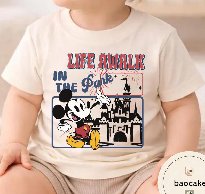 May include: Cream-coloured t-shirt featuring a graphic of Mickey Mouse with the text "LIFE A WALK IN THE Park". The design includes a castle silhouette and fireworks. The shirt is likely made of soft fabric.