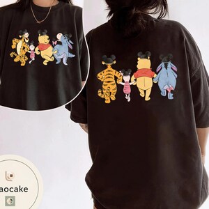 May include: Black t-shirt featuring a cartoon graphic of Winnie the Pooh, Tigger, Piglet, and Eeyore. The front shows the characters in a line, and the back shows the same characters. The characters are wearing Mickey Mouse ears.
