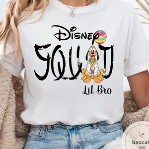 Puede incluir: Camiseta blanca con un dise&ntilde;o negro que dice "Disney Squad" con una imagen de dibujos animados de Pluto sosteniendo un globo arco&iacute;ris. Las palabras "Lil Bro" est&aacute;n impresas debajo.