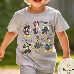 May include: A light gray t-shirt featuring a colorful graphic of Disney characters in graduation attire. Mickey Mouse, Minnie Mouse, Donald Duck, Goofy, and Pluto are depicted with graduation caps and gowns. The shirt has the text "Mickey Mouse" and character names.