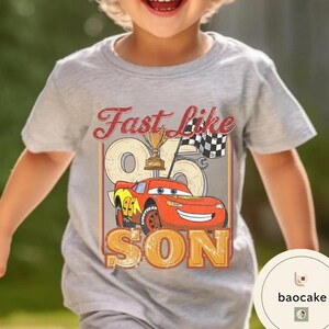 May include: A light gray t-shirt featuring a graphic of a red race car with the text "Fast Like 95 Son". The design includes a trophy and checkered flag, with a vintage, distressed style.