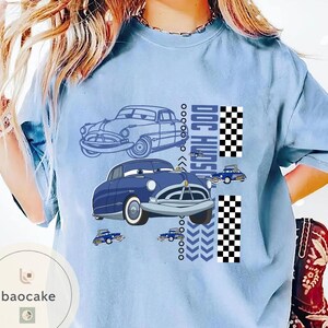 May include: Pale blue t-shirt with a cartoon car design. The design features two blue cars, the text "DOC HUDSON", and a checkered flag pattern. Small car graphics and chevron arrows are also included.