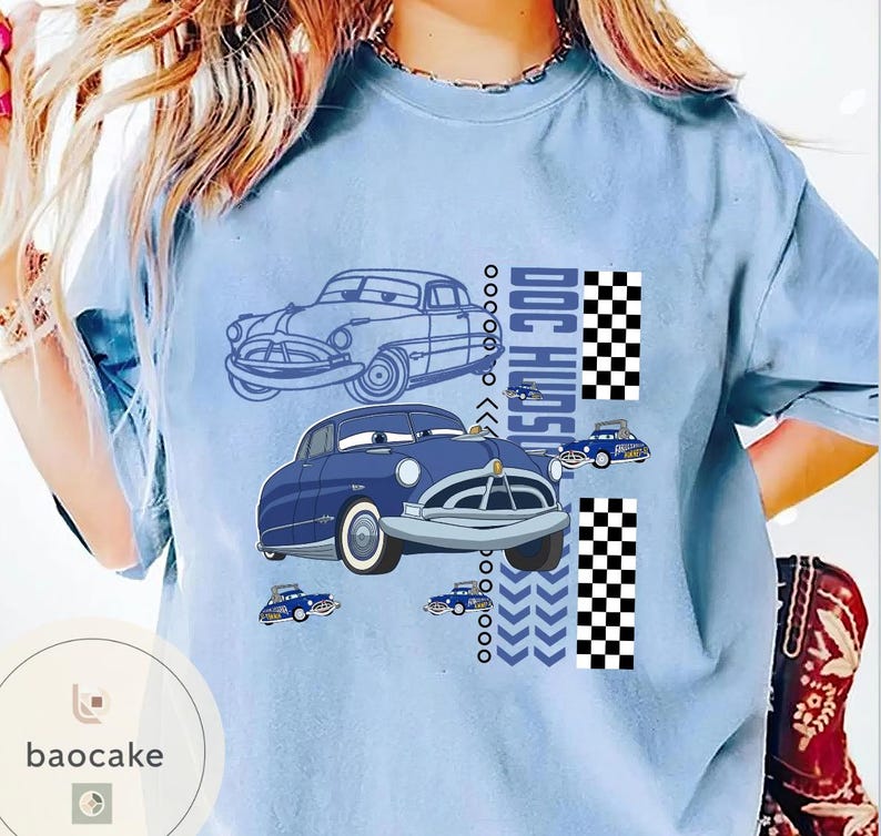 May include: Pale blue t-shirt with a cartoon car design. The design features two blue cars, the text "DOC HUDSON", and a checkered flag pattern. Small car graphics and chevron arrows are also included.