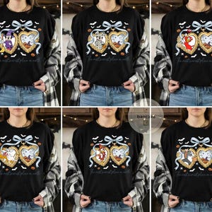 May include: Black long-sleeved top with a Halloween graphic. The design features a heart-shaped locket with cartoon characters, pumpkins, bats, and the text "The most scariest place on earth". A light blue bow and ribbon complete the design.