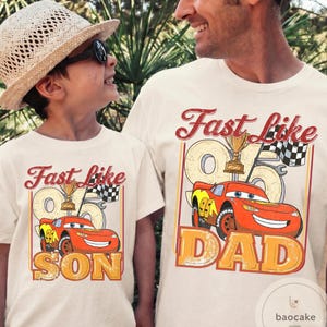 May include: Two matching cream-colored t-shirts with the text "Fast Like" above a cartoon race car and the text "SON" and "DAD" below. The race car is red and yellow with the number 95. A trophy and checkered flag are also featured.