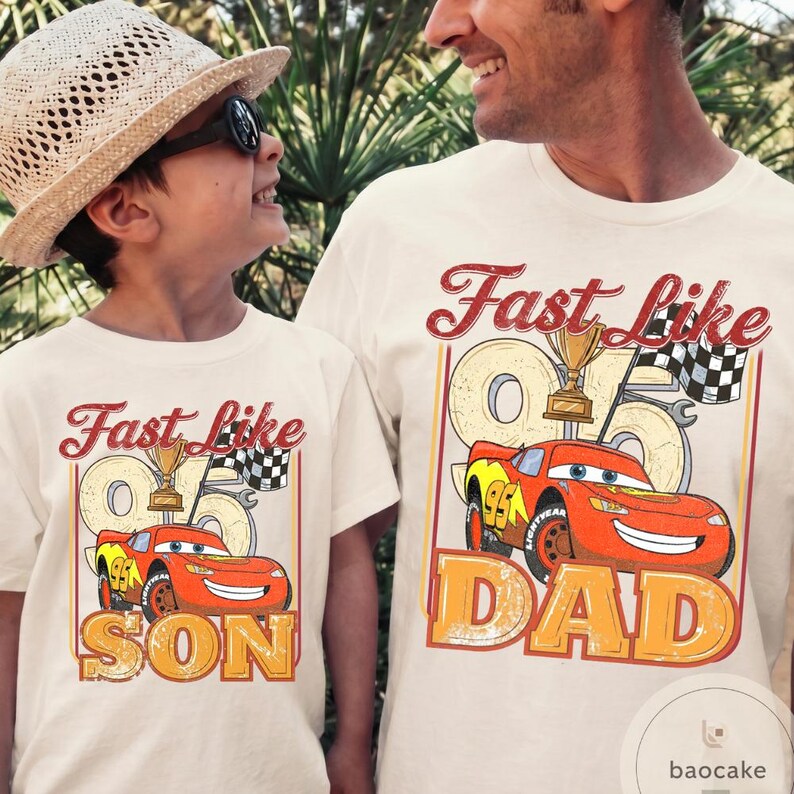 May include: Two matching cream-colored t-shirts with the text "Fast Like" above a cartoon race car and the text "SON" and "DAD" below. The race car is red and yellow with the number 95. A trophy and checkered flag are also featured.