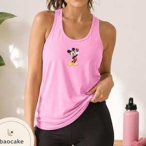 May include: A light pink racerback tank top featuring a small graphic of Mickey Mouse in the center. The character is depicted in classic colors, wearing red shorts and yellow shoes, with one hand raised. The top has a scoop neckline and sleeveless design.