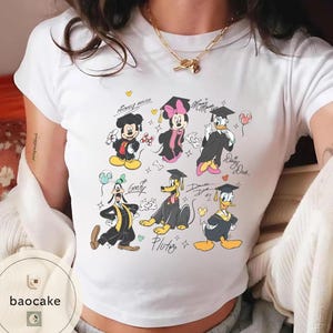May include: White t-shirt featuring cartoon characters Mickey Mouse, Minnie Mouse, Donald Duck, Daisy Duck, Goofy, and Pluto in graduation attire. The characters are wearing graduation caps and gowns, with some holding balloons. The shirt has the text "Mickey Mouse", "Minnie Mouse", "Daisy Duck", and "Pluto".
