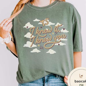 K&ouml;nnte beinhalten: Ein olivgr&uuml;nes Kurzarm-T-Shirt mit einem Grafikdruck aus wei&szlig;en Wolken, kleinen dunklen Herzen und dem Text TS I knew it, I knew you in einer seilartigen Schriftart. Das T-Shirt hat einen Rundhalsausschnitt und eine lockere Passform.