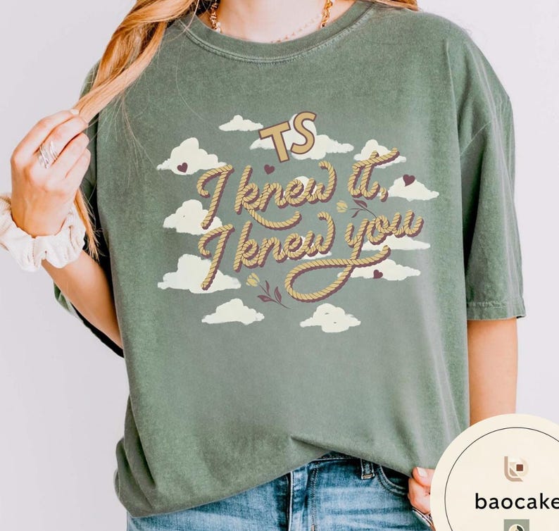 K&ouml;nnte beinhalten: Ein olivgr&uuml;nes Kurzarm-T-Shirt mit einem Grafikdruck aus wei&szlig;en Wolken, kleinen dunklen Herzen und dem Text TS I knew it, I knew you in einer seilartigen Schriftart. Das T-Shirt hat einen Rundhalsausschnitt und eine lockere Passform.