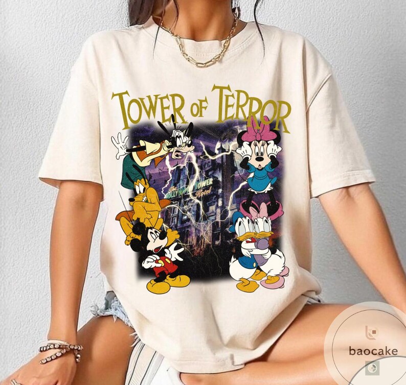 May include: Cream-colored t-shirt featuring the text "Tower of Terror" in gold lettering. The graphic includes cartoon characters Mickey Mouse, Minnie Mouse, Donald Duck, Goofy, and Pluto in front of a purple and blue background.