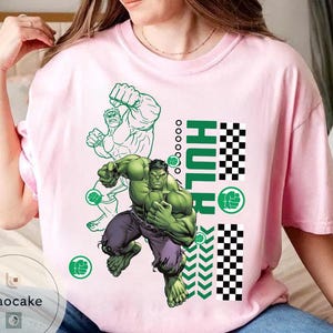 May include: A light pink t-shirt featuring a graphic of the Hulk in a dynamic pose. The design includes the word "HULK" in green, a green outline of the Hulk, and a checkered pattern. The Hulk is depicted in green with purple pants.