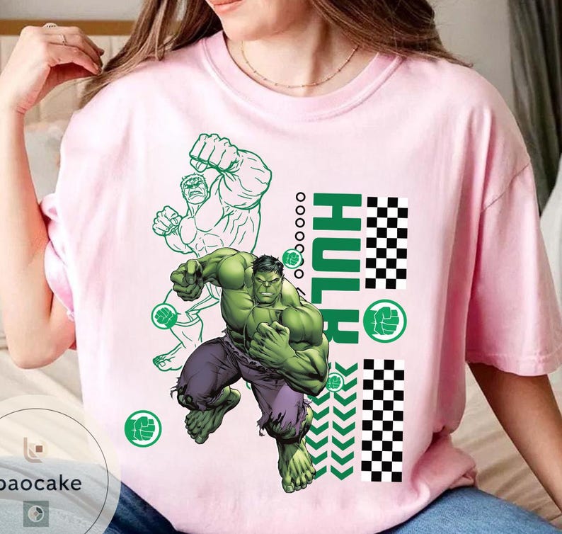 May include: A light pink t-shirt featuring a graphic of the Hulk in a dynamic pose. The design includes the word "HULK" in green, a green outline of the Hulk, and a checkered pattern. The Hulk is depicted in green with purple pants.