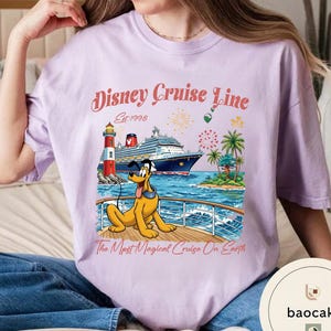 K&ouml;nnte beinhalten: Helllila T-Shirt mit einem Aufdruck von Pluto auf einem Kreuzfahrtschiff, einem Leuchtturm und Palmen. Der Text "Disney Cruise Line Est. 1998" und "The Most Magical Cruise On Earth" sind ebenfalls abgebildet.