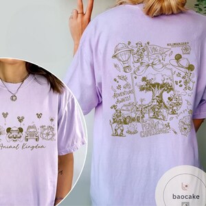 May include: A light purple t-shirt featuring a detailed graphic design. The front of the shirt says "Animal Kingdom" with Mickey Mouse and other park-themed icons. The back of the shirt has a map-like design with the words "Adventure Awaits!" and "Kilimanjaro".