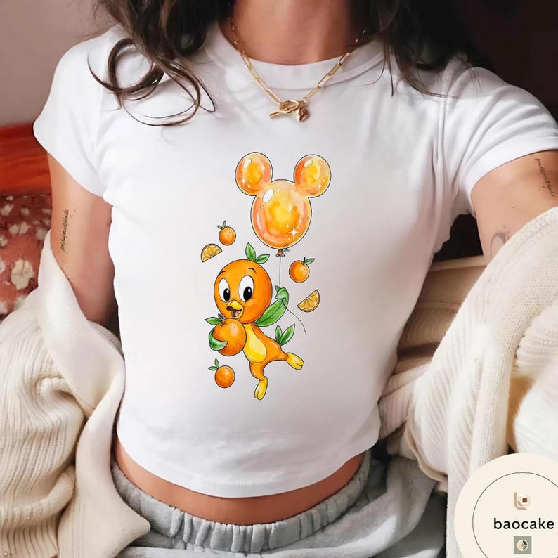 Op de afbeelding: Wit T-shirt met een cartoon oranje vogel die een sinaasappel vasthoudt, vergezeld van een Mickey Mouse-vormige ballon. Het ontwerp bevat sinaasappelschijfjes en kleine sinaasappels. De vogel heeft groene bladeren en een vrolijke uitdrukking.
