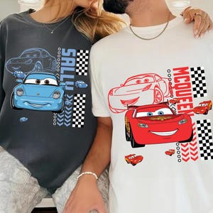 May include: Two t-shirts with cartoon car designs. One is dark grey with a blue car and the name "SALLY". The other is white with a red car and the name "MCQUEEN". Both shirts feature a chequered flag design.