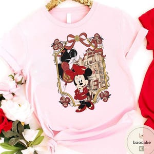 May include: A light pink t-shirt featuring a Minnie Mouse graphic. The graphic includes Minnie Mouse in a red outfit, a building, and a balloon that says "Hi". The design is framed with a gold border and red roses.