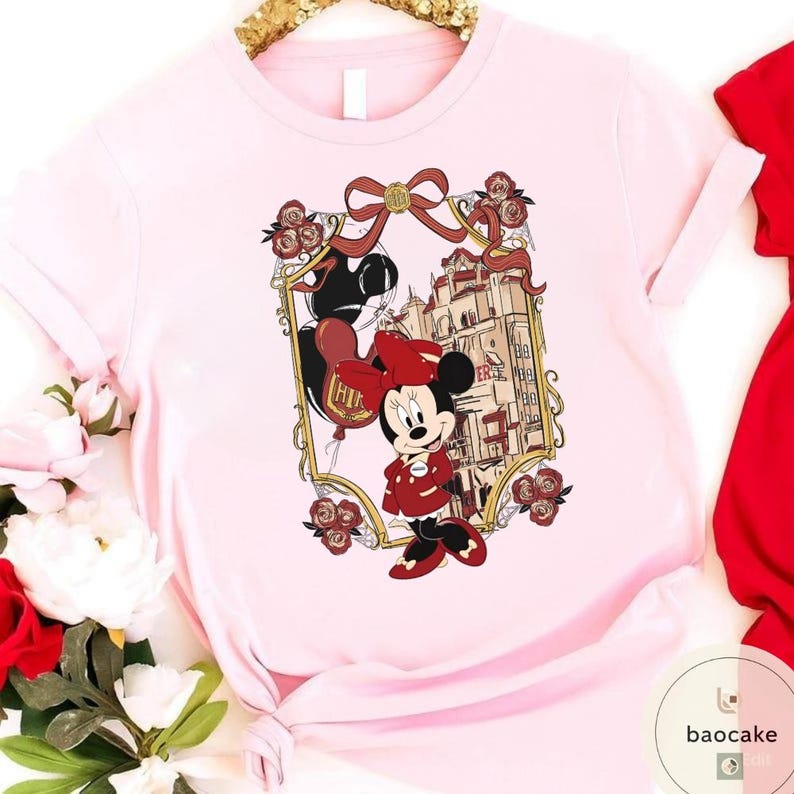 May include: A light pink t-shirt featuring a Minnie Mouse graphic. The graphic includes Minnie Mouse in a red outfit, a building, and a balloon that says "Hi". The design is framed with a gold border and red roses.
