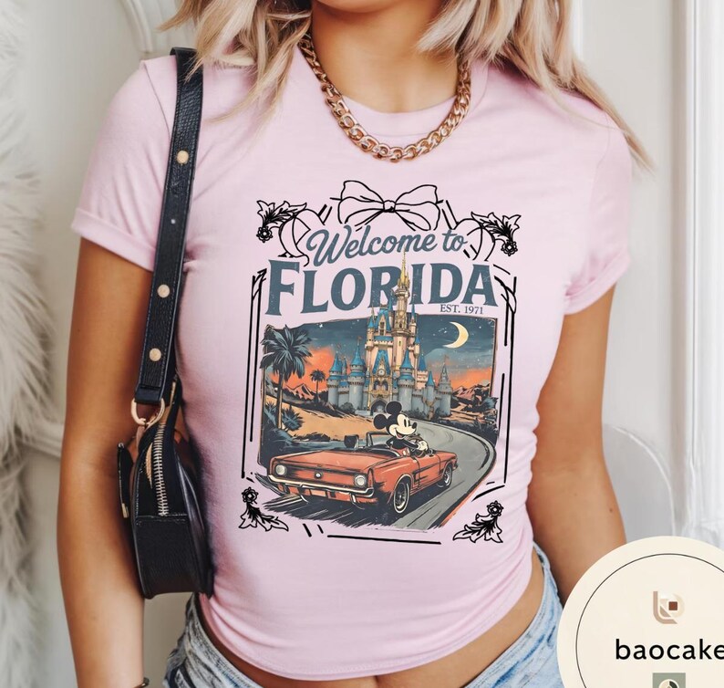 May include: A light pink t-shirt featuring a graphic that says "Welcome to Florida" with a vintage car driven by Mickey Mouse in front of a castle. The graphic is framed with decorative elements.