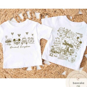 May include: Two white t-shirts with gold-colored graphics. One shirt says "Animal Kingdom" with Mickey Mouse and other park icons. The other shirt says "Adventure Awaits!" with a safari theme and "Kilimanjaro" text.