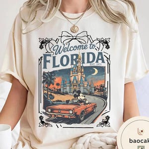 May include: Cream-colored t-shirt featuring a graphic of Mickey Mouse driving a red convertible towards a castle. The text "Welcome to Florida" and "EST. 1971" are displayed above the image in a decorative frame.