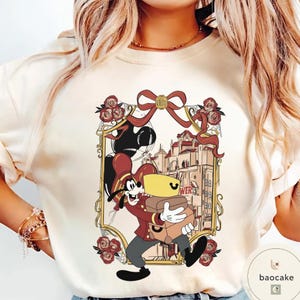 May include: Cream-colored sweatshirt featuring a cartoon illustration of Goofy in a bellhop uniform carrying luggage. The design is framed with a gold border, red bow, and roses. The word "WER" is visible.