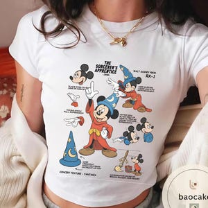 May include: White t-shirt featuring a graphic of Mickey Mouse from "The Sorcerer's Apprentice" (1940). The design includes various poses of Mickey in his sorcerer attire, with text annotations. The shirt has a crew neck.