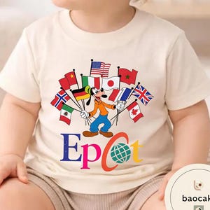 May include: Cream-coloured t-shirt featuring a cartoon of Goofy holding flags from various countries. The word "Epcot" is printed in colourful letters below the cartoon, with a globe graphic. The shirt is likely for children.