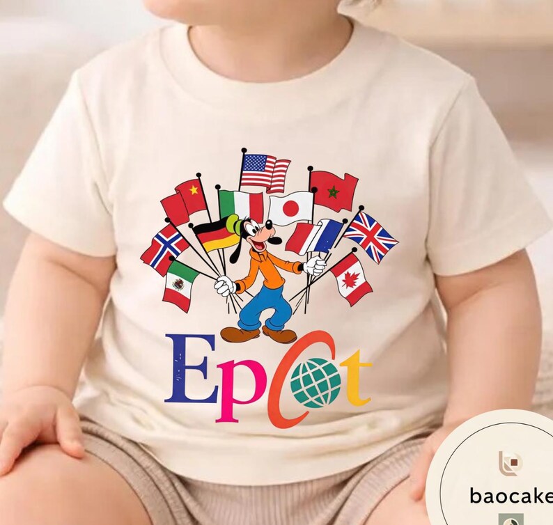 May include: Cream-coloured t-shirt featuring a cartoon of Goofy holding flags from various countries. The word "Epcot" is printed in colourful letters below the cartoon, with a globe graphic. The shirt is likely for children.