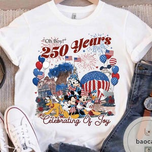 Puede incluir: Camiseta blanca con un gr&aacute;fico colorido que celebra 250 a&ntilde;os. El dise&ntilde;o presenta a Mickey Mouse, Minnie Mouse, Donald Duck y Pluto con atuendo patri&oacute;tico, junto con fuegos artificiales, globos y el texto "Oh Boy!" y "Celebrating Of Joy."