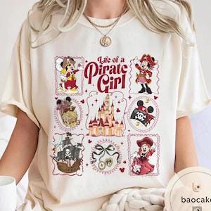 May include: Cream-colored t-shirt featuring a "Life of a Pirate Girl" design. The design includes cartoon illustrations of a pirate-themed character, a treasure chest, a pirate ship, and a castle, all framed in decorative borders.