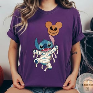 May include: Purple t-shirt featuring a Halloween-themed graphic. The design includes a blue cartoon character wrapped in bandages, holding a jack-o'-lantern balloon. The jack-o'-lantern is brown with a carved face. The shirt has short sleeves.